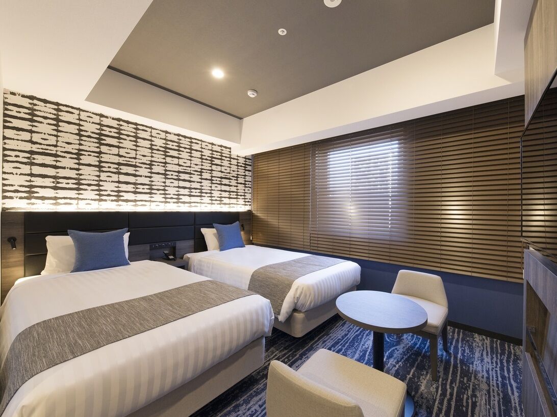 Une chambre d’hôtel avec deux lits, une petite table ronde, une chaise, un téléviseur et un mur décoré d’un motif.
