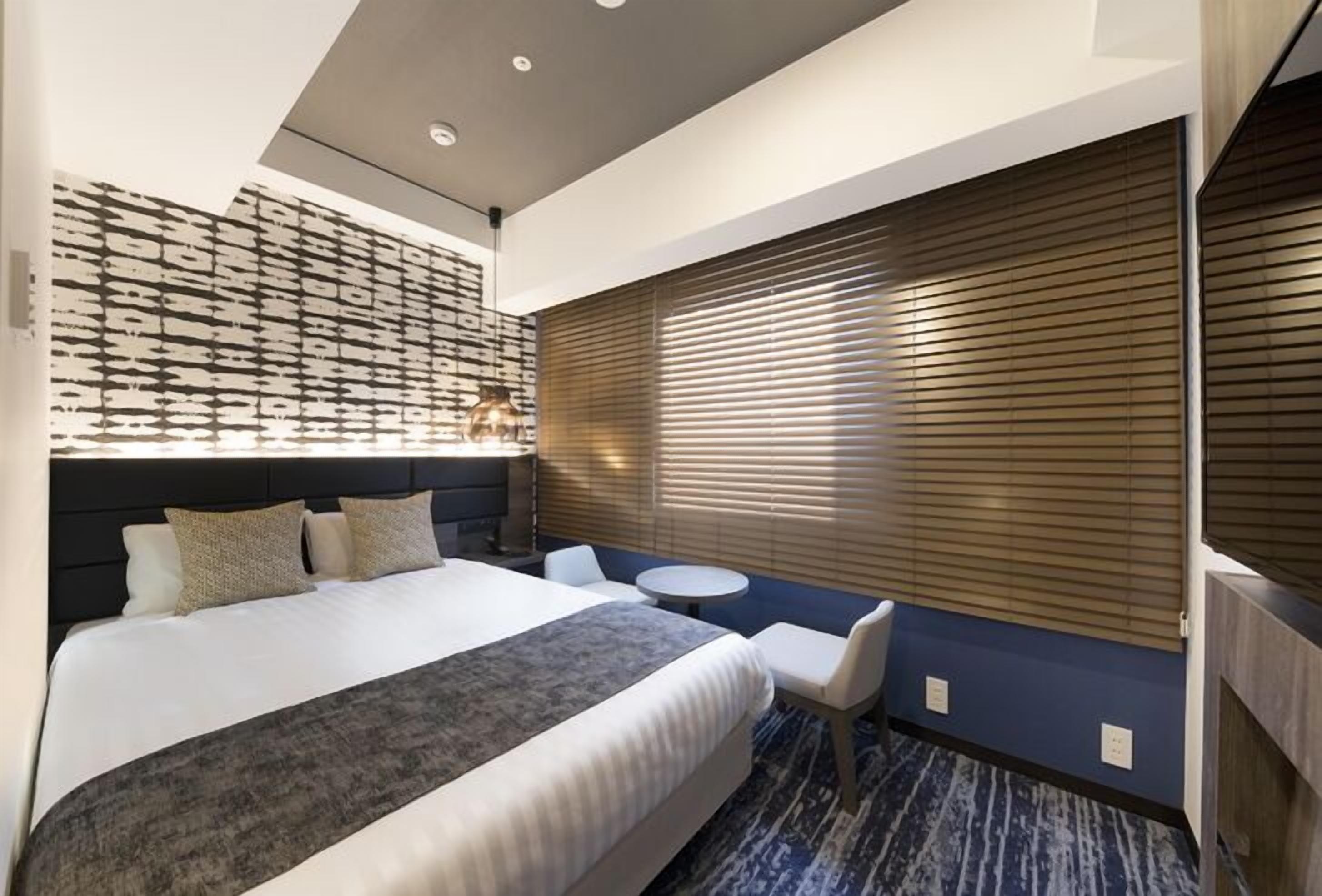 Une chambre d’hôtel avec un lit, une petite table ronde, une chaise, un t éléviseur fixé au mur et une fenêtre avec des stores.
