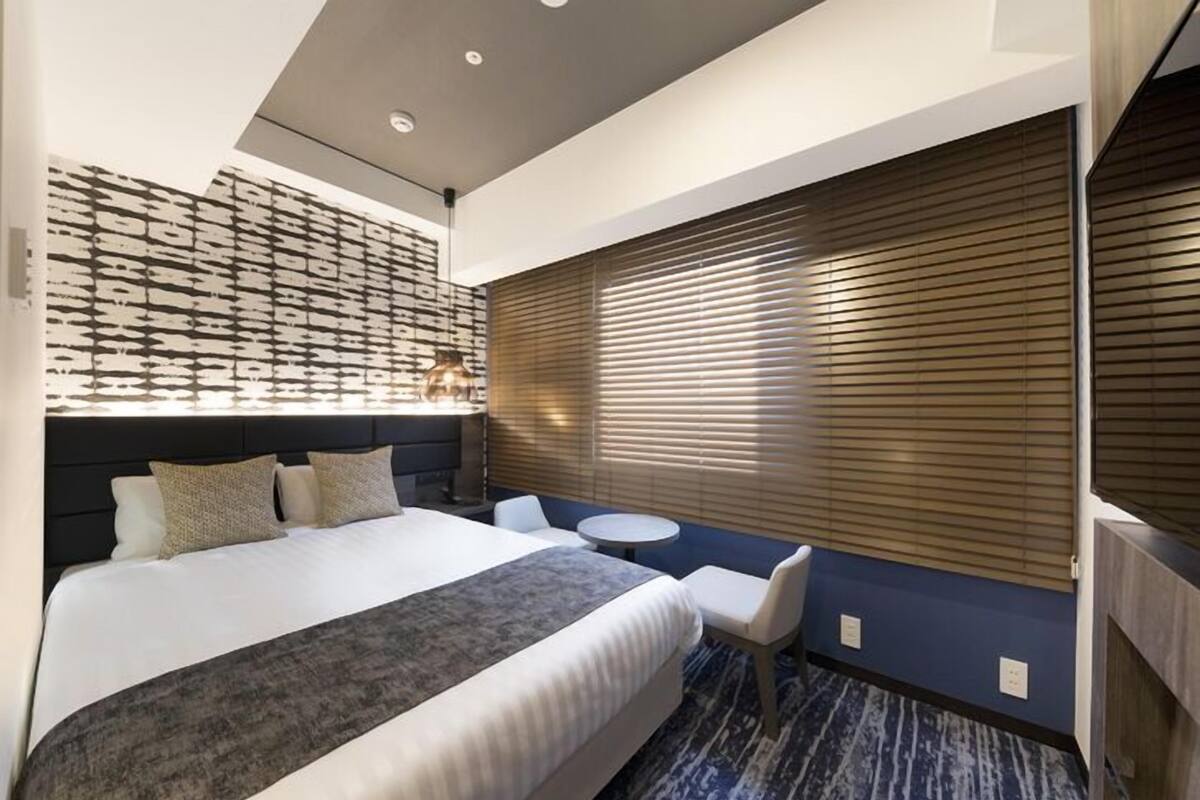 Ein Hotelzimmer mit Bett, kleinem runden Tisch, Stuhl, Wand-Fernseher und Fenster mit Jalousien.
