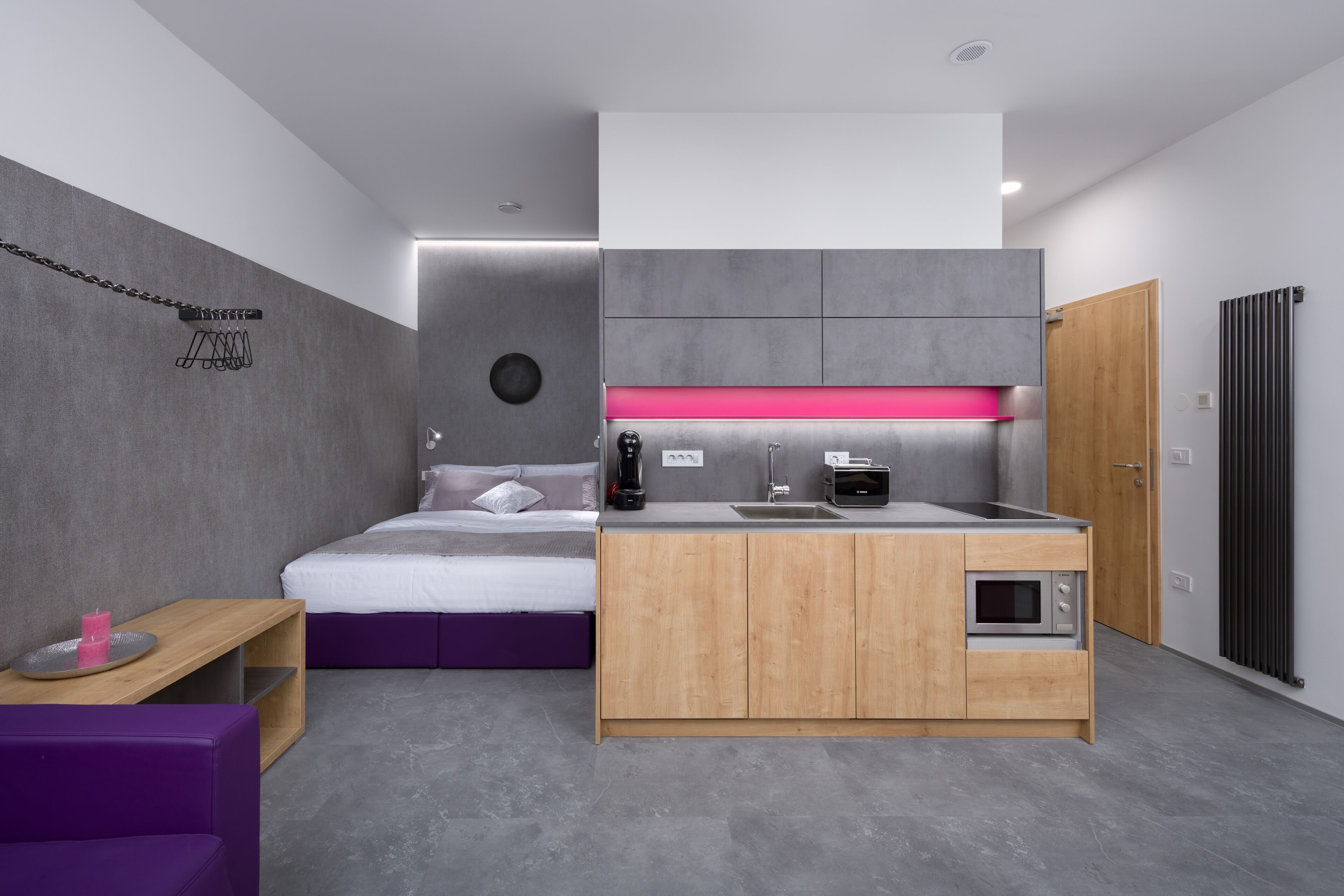 A modern hotel room with a bed, a small kitchenette, a wooden cabinet, and a purple sofa.