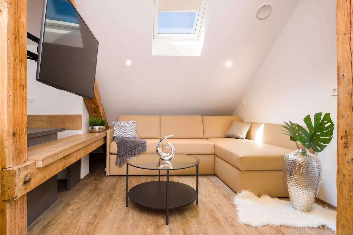 Een moderne woonkamer met een glazen salontafel, een beige bank, een houten bankje, een aan een houten balk bevestigde televisie en een dakraam.