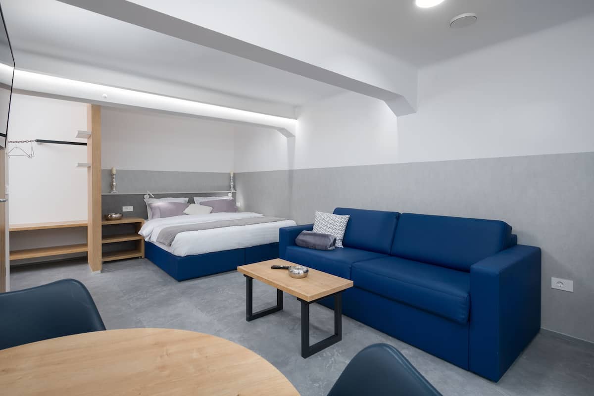 Een moderne, compacte kamer met een blauwe bank, een klein houten tafeltje, een bed met een wit en grijs hoofdeinde, en een inbouwkast.