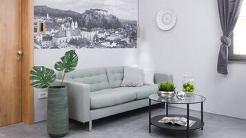 A modern living room with a grey sofa, a round glass coffee table, a large potted plant, and a wall-mounted picture.