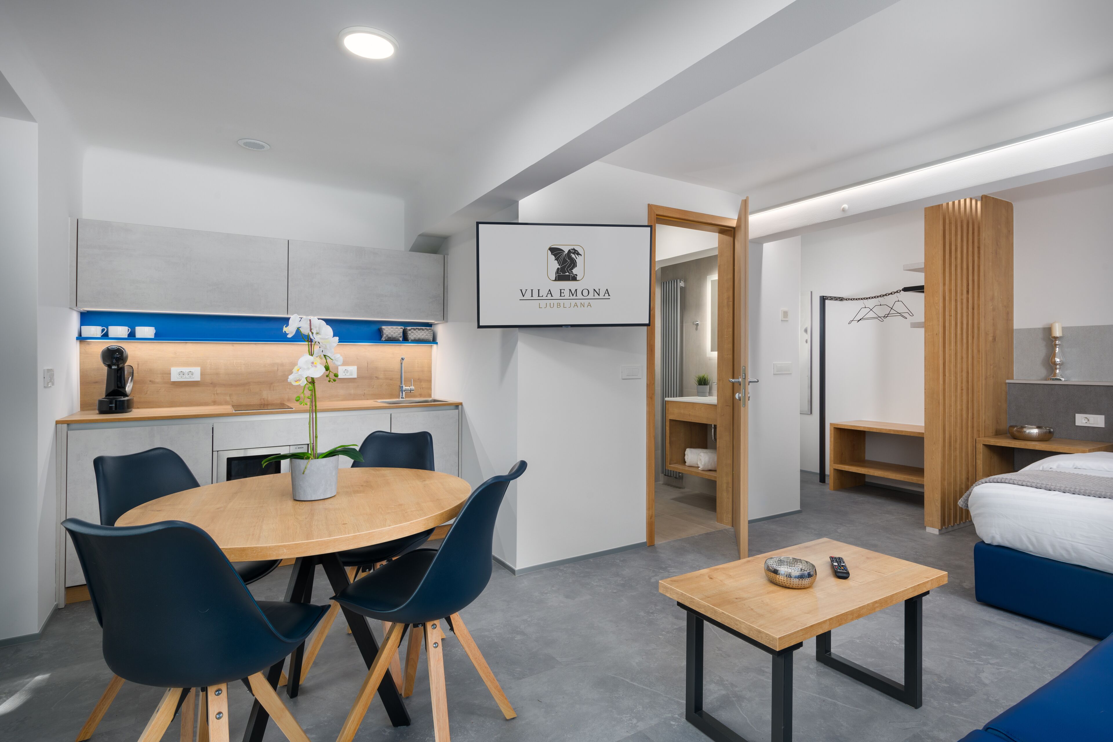 Ein modernes Hotelzimmer mit einem Holztisch, blauen Stühlen, einem kleinen Beistelltisch aus Holz, einem Bett mit blauer Bettwäsche, einem Holzschrank und einem gerahmten Logo an der Wand.