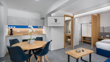 Ein modernes Hotelzimmer mit einem Holztisch, blauen Stühlen, einem kleinen Beistelltisch aus Holz, einem Bett mit blauer Bettwäsche, einem Holzschrank und einem gerahmten Logo an der Wand.
