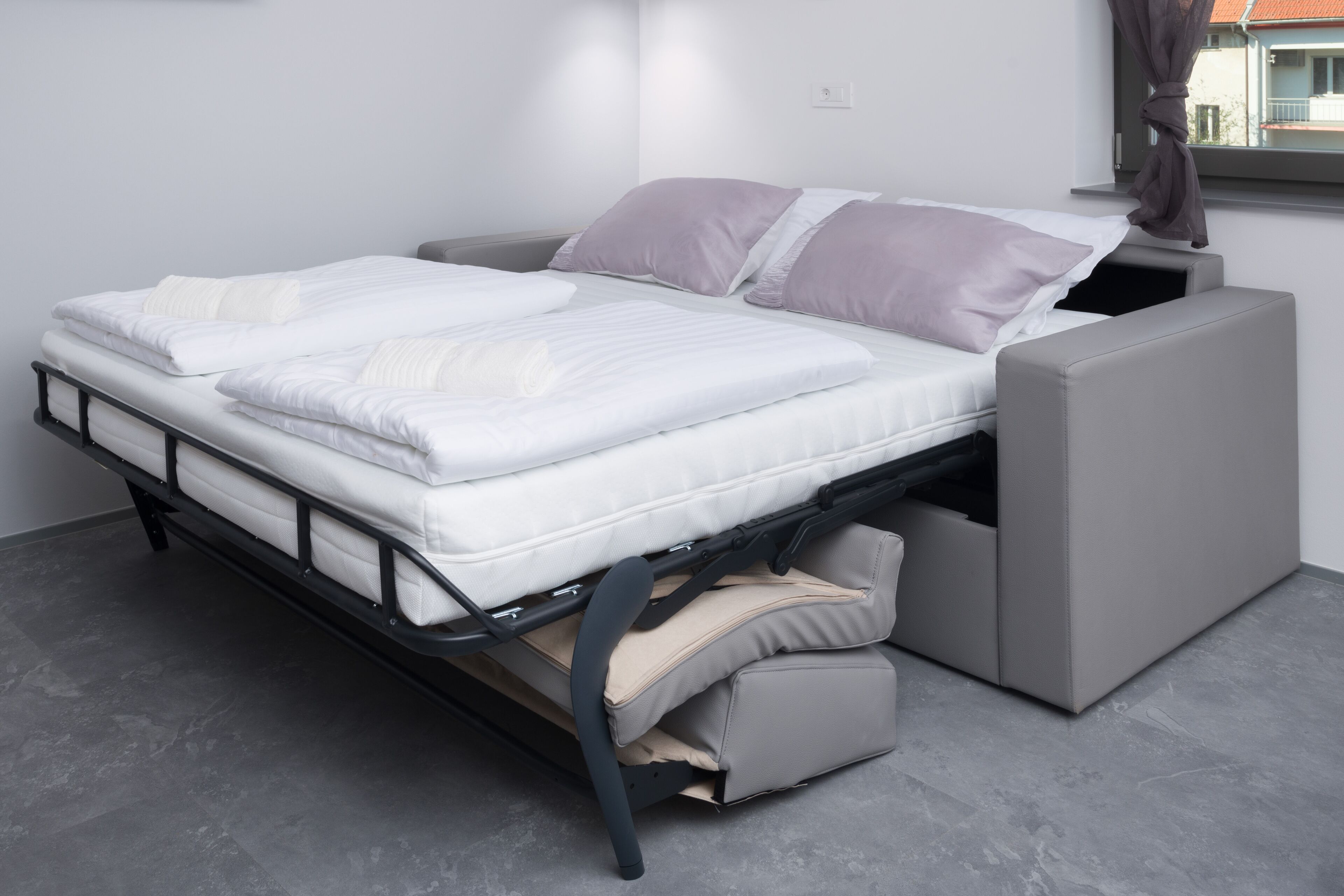 A bed with a grey mattress and white bedding, a grey upholstered bench, and a window with curtains.
