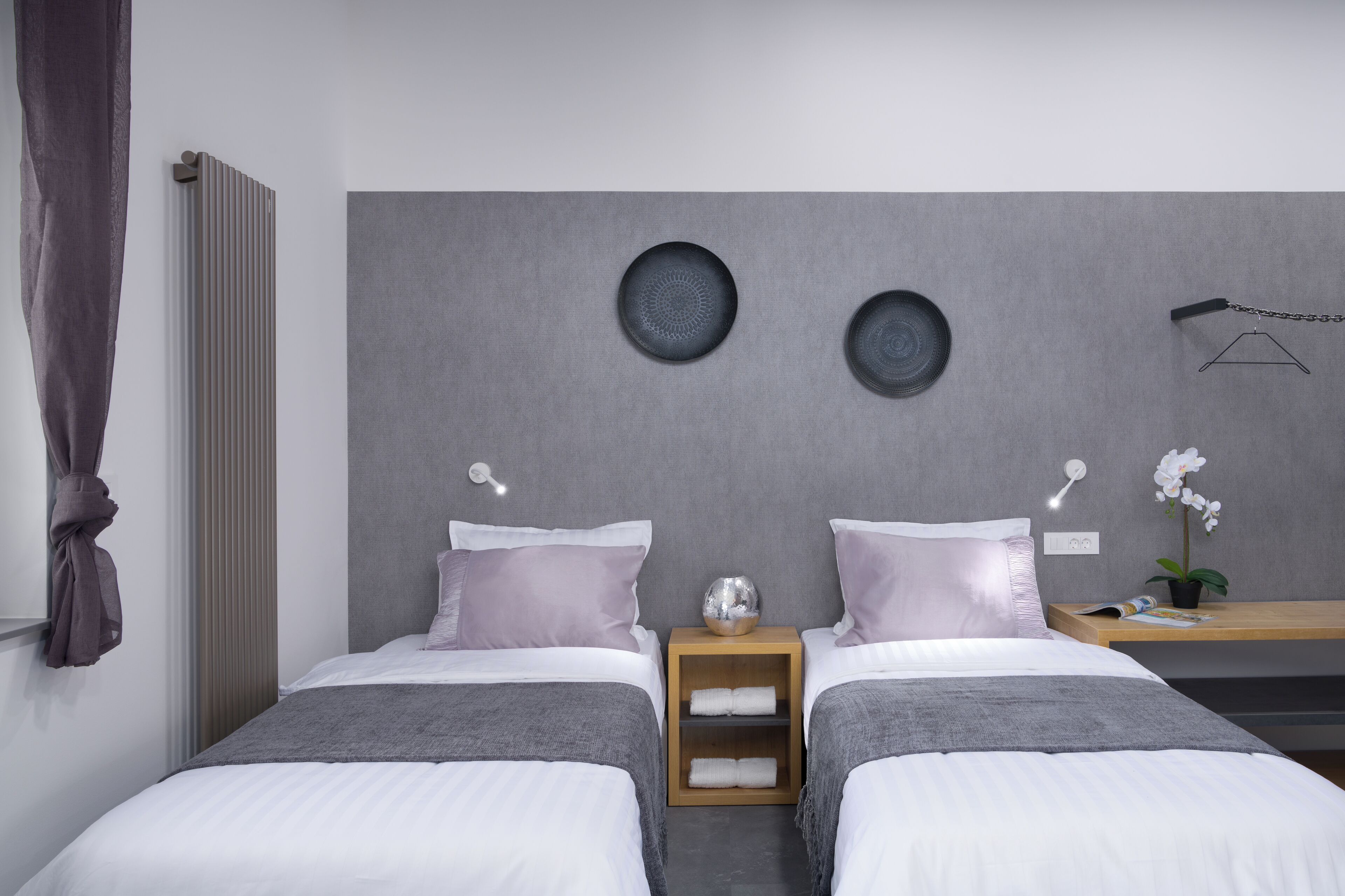 Una habitación de hotel moderna con dos camas, una mesita de noche de madera, una pequeña mesa y una lámpara fijada a la pared.