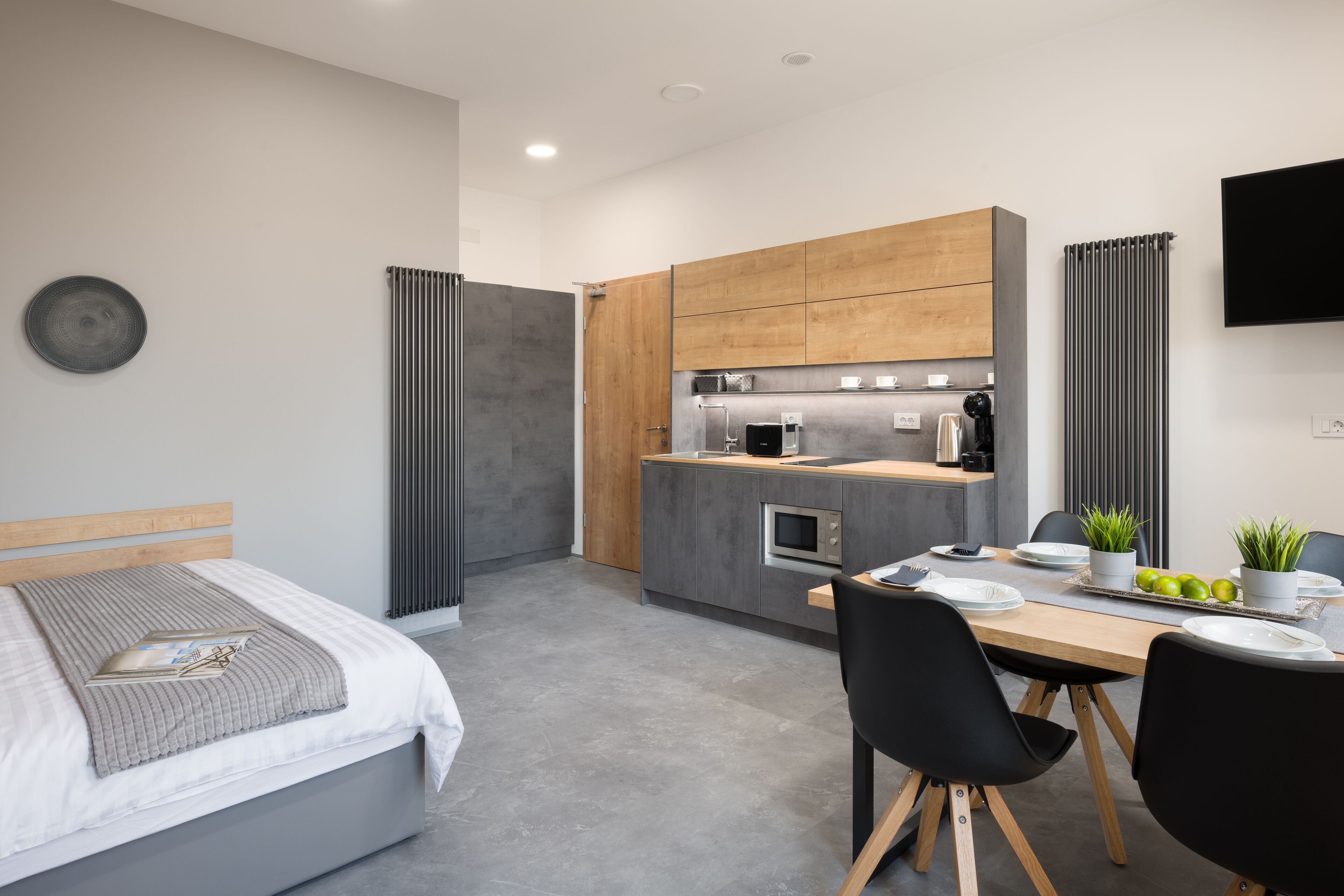 A modern hotel room with a bed, bedside table, and a small kitchenette.