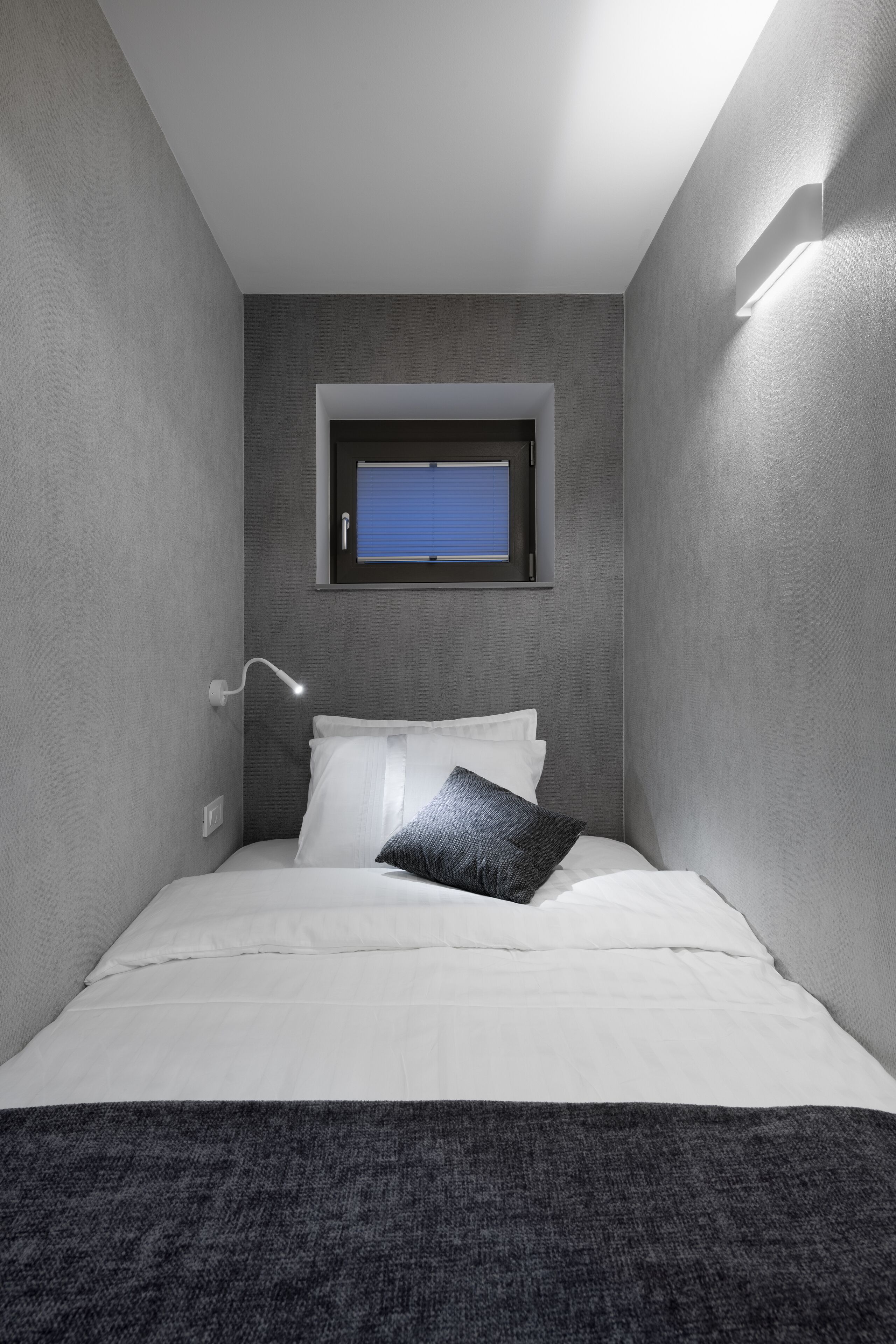 A small, modern bedroom with a single bed, a window, and a wall-mounted light fixture.