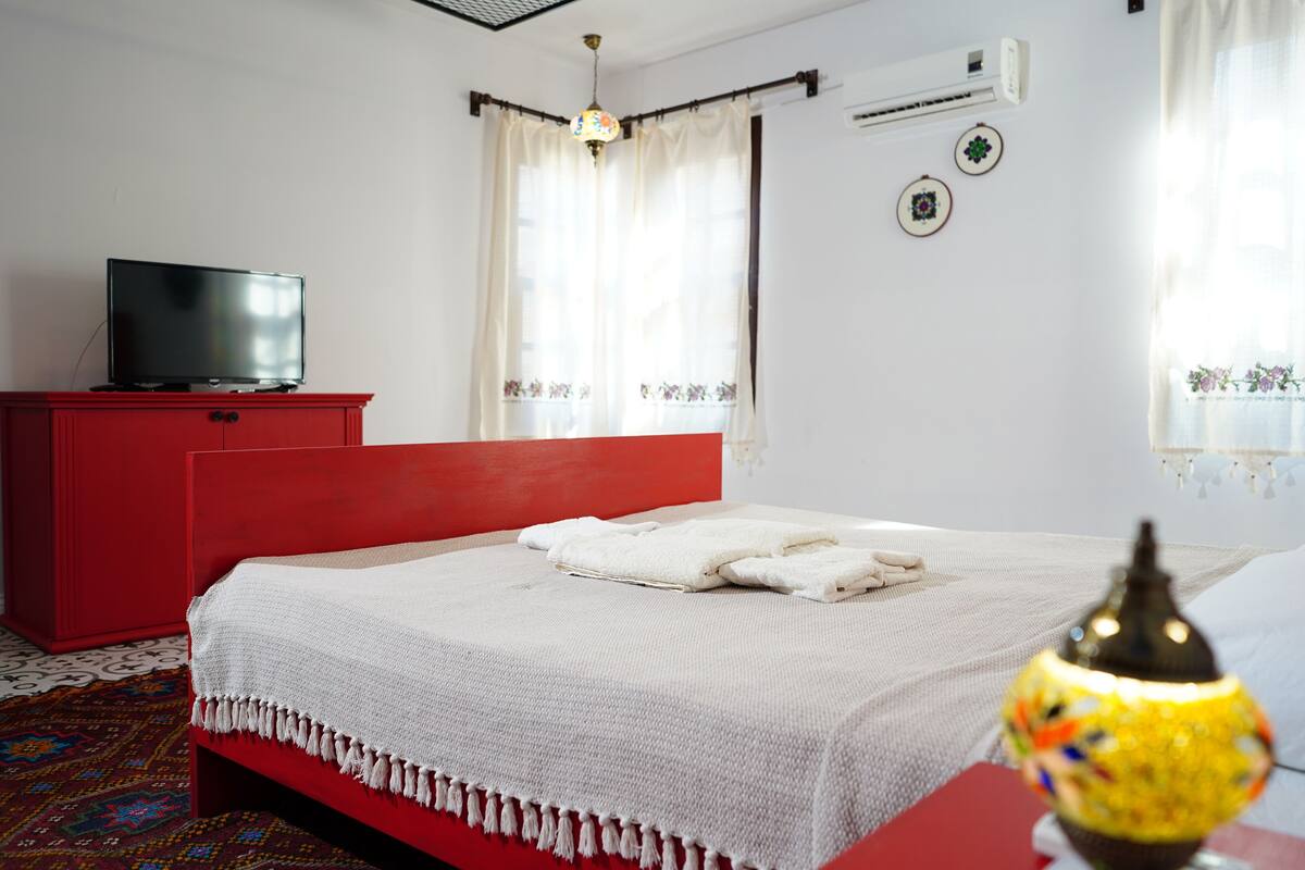 A room with a red bed, white bedding, a television on a red cabinet, and a decorative lamp.