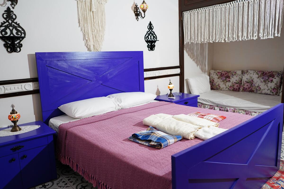 A bedroom with a blue bed, white pillows, a pink blanket, and a nightstand with a lamp.