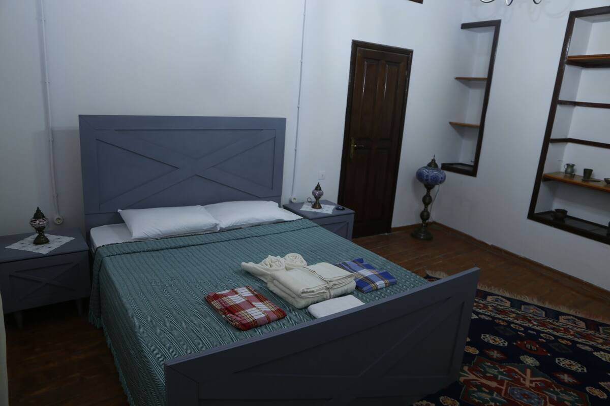 A neatly made bed with a blue headboard, a bedside table with a lamp, and a wall-mounted shelf.