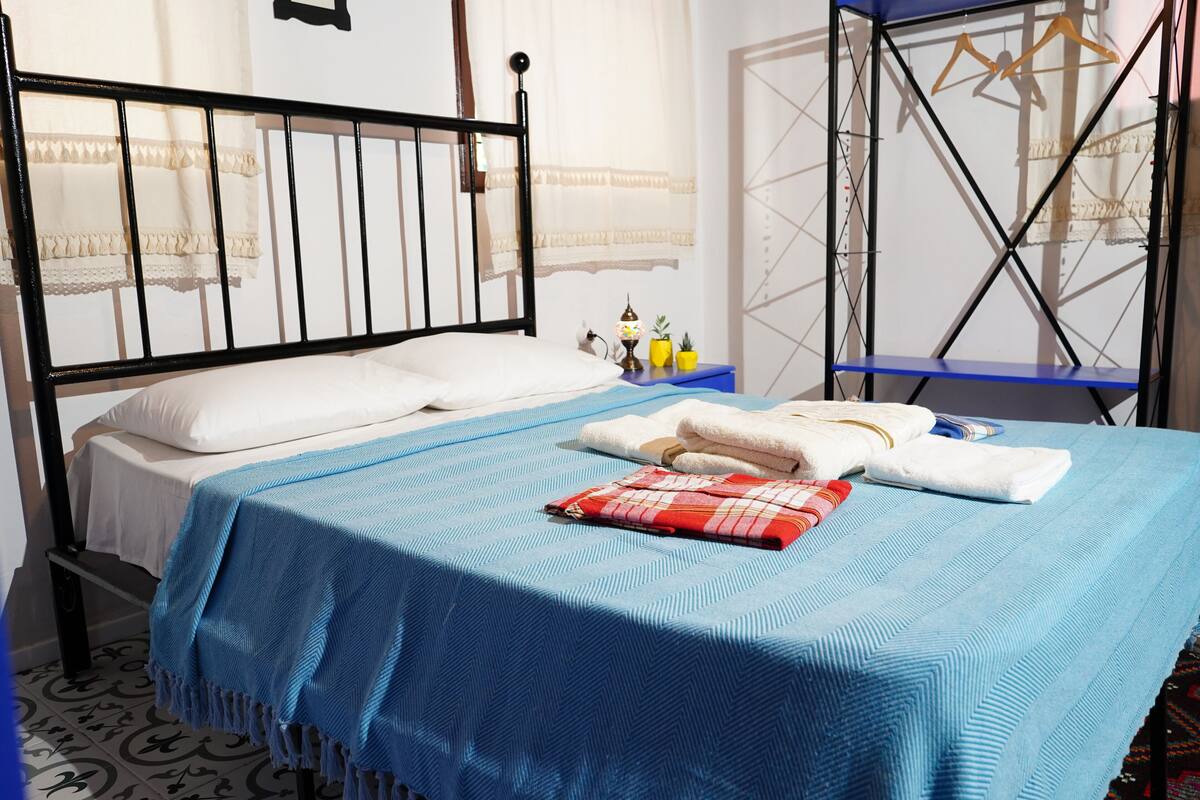 A neatly made bed with a blue bedspread and white pillows, a small bedside table with a lamp and two yellow cups, and a blue shelf unit against the wall.