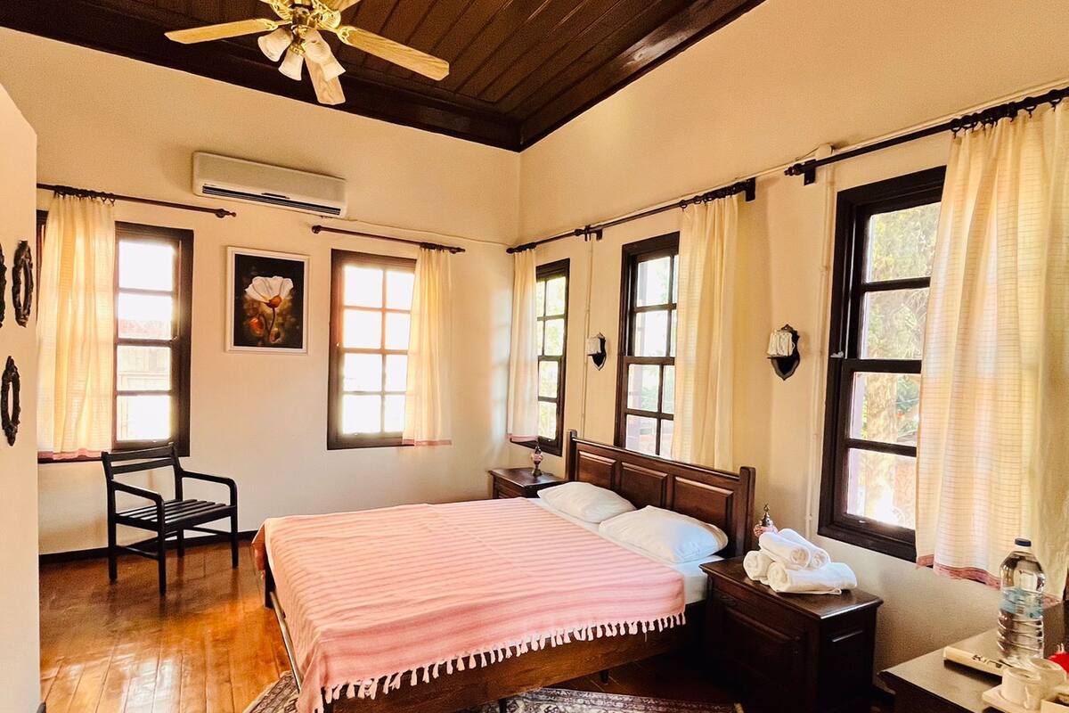 A bedroom with a wooden ceiling, a bed with a pink bedspread, a patterned rug, a desk with a lamp, and a nightstand with a water bottle.