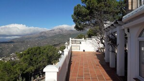 Um terraço com vista para uma paisagem montanhosa, colunas brancas e piso de azulejos.