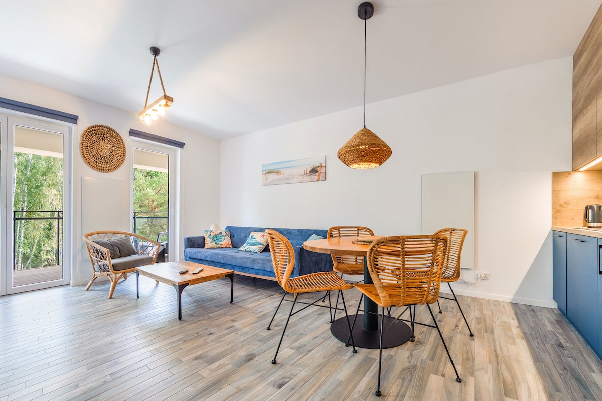 Een moderne woonkamer met een blauwe bank, rieten stoelen, een houten salontafel en een eettafel. Er is een groot raam met uitzicht op bomen en een balkon.