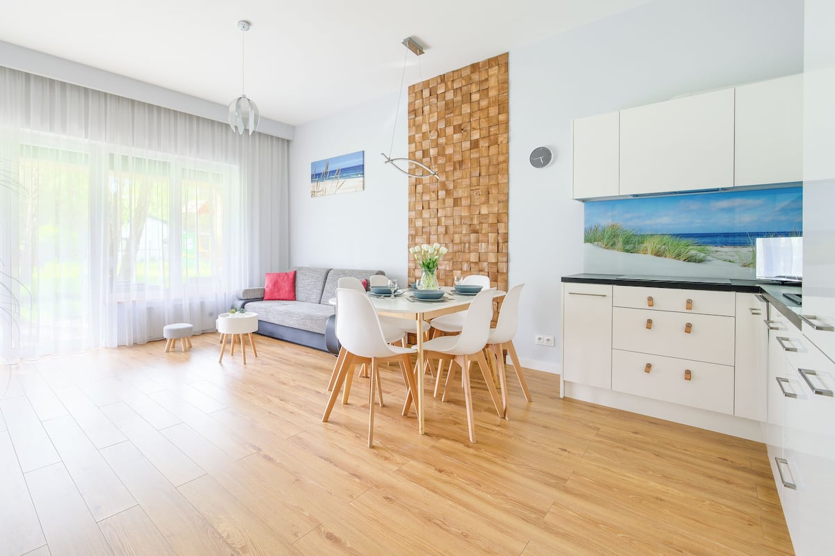 Een moderne woonkamer met een eethoek, houten vloeren, een groot raam met lichte gordijnen, een grijze bank, een houten accentmuur en een keuken met witte kasten.