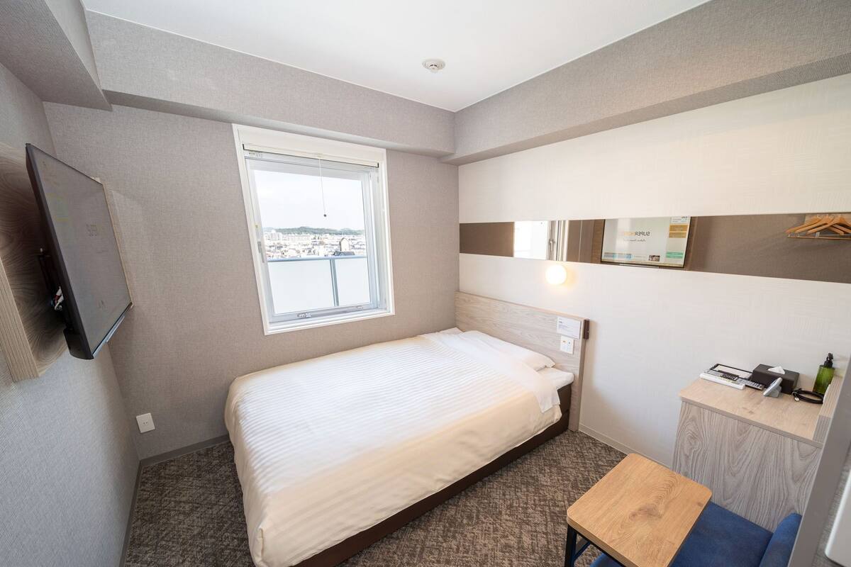 A hotel room with a single bed, a small desk, a mirror, and a window with a city view.