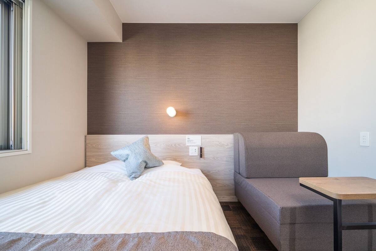 A modern hotel room with a bed, a sofa, a small table, and a wall-mounted light.