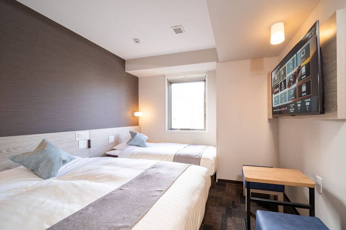 A hotel room with two beds, a desk, a window, and a mounted TV.