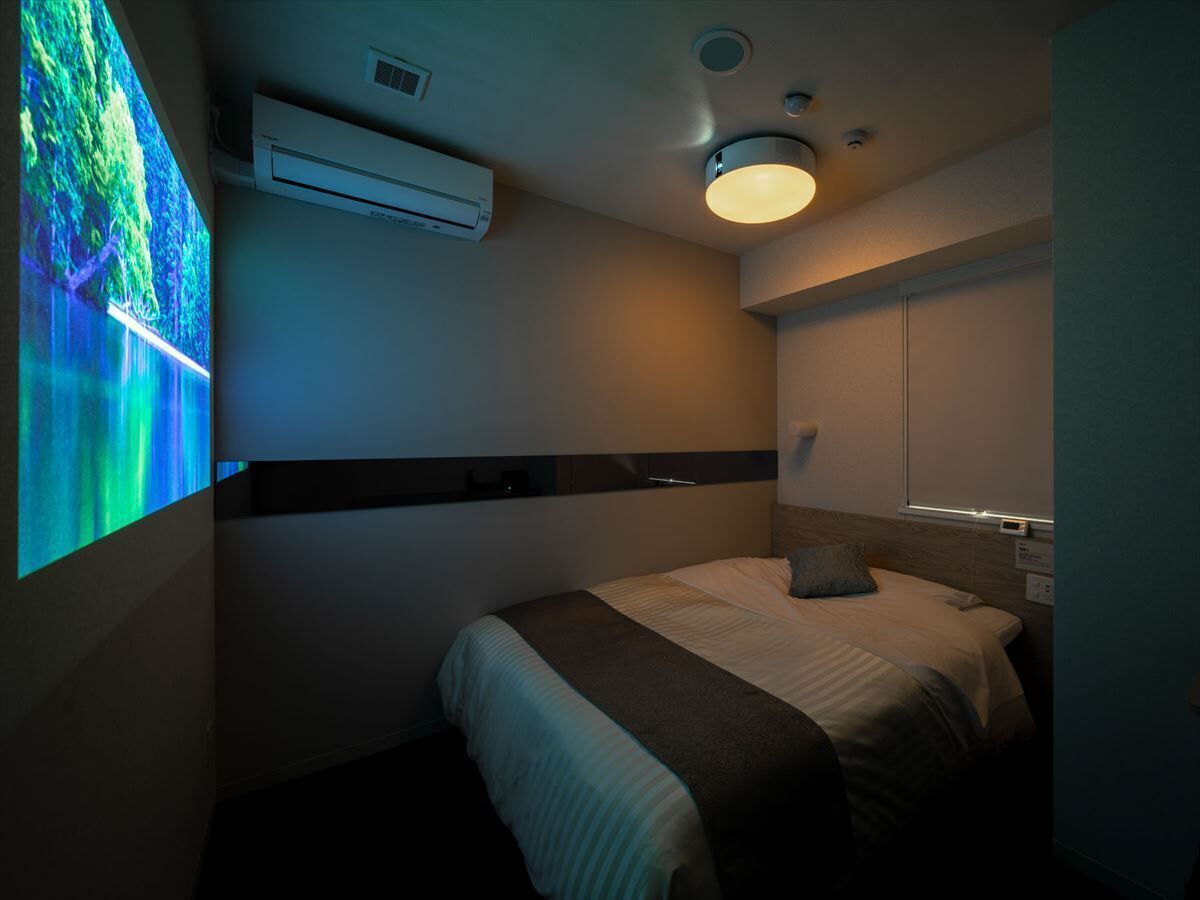 Une chambre d’hôtel équipée d’un lit, d’un climatiseur fixé au mur, d’un luminaire au plafond et d’un écran de projection diffusant une scène de la nature.
