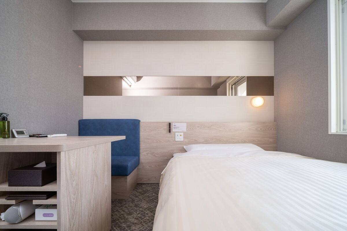 A compact hotel room with a bed, a desk, a chair, a mirror, and a small shelf with items.