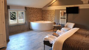 Un dormitorio moderno con una cama grande, una zona de estar con una silla, una mesita pequeña, una bañera y un televisor montado en la pared.