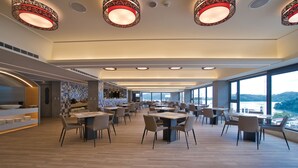 Un amplio comedor con múltiples mesas y sillas, ventanas grandes con vistas a la ciudad e iluminación decorativa en el techo.