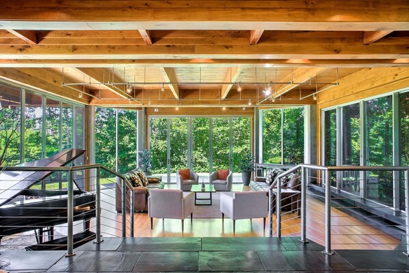 Amplia sala de estar con un piano de cola, muebles modernos y grandes ventanas que ofrecen vista al bosque.