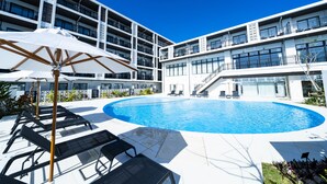 Un hôtel moderne doté d’une piscine, de chaises longues et d’un espace abrité avec des parasols.
