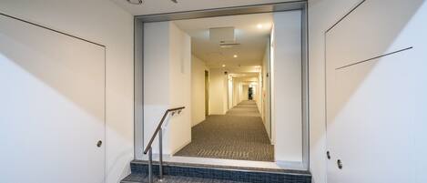 A modern hotel corridor with a carpeted staircase, a metal handrail, and a view into a long hallway with doors on either side.