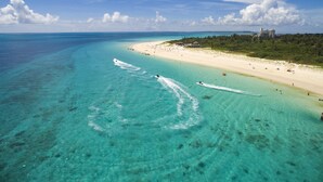 Une plage aux eaux turquoise et limpides, un rivage sablonneux et quelques bâtiments visibles au loin.