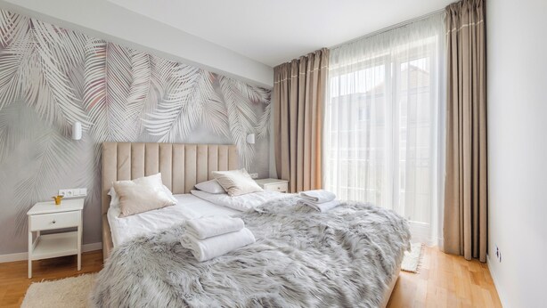 A modern bedroom with a large bed, a nightstand, a wall-mounted lamp, and sheer curtains. - Apartamenty Nadmorskie Sun & Snow (Sopot)