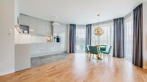 A modern kitchen with white cabinets, a dining area with a round table and teal chairs, and large windows with grey curtains. - Apartamenty Nadmorskie Sun & Snow (Sopot)