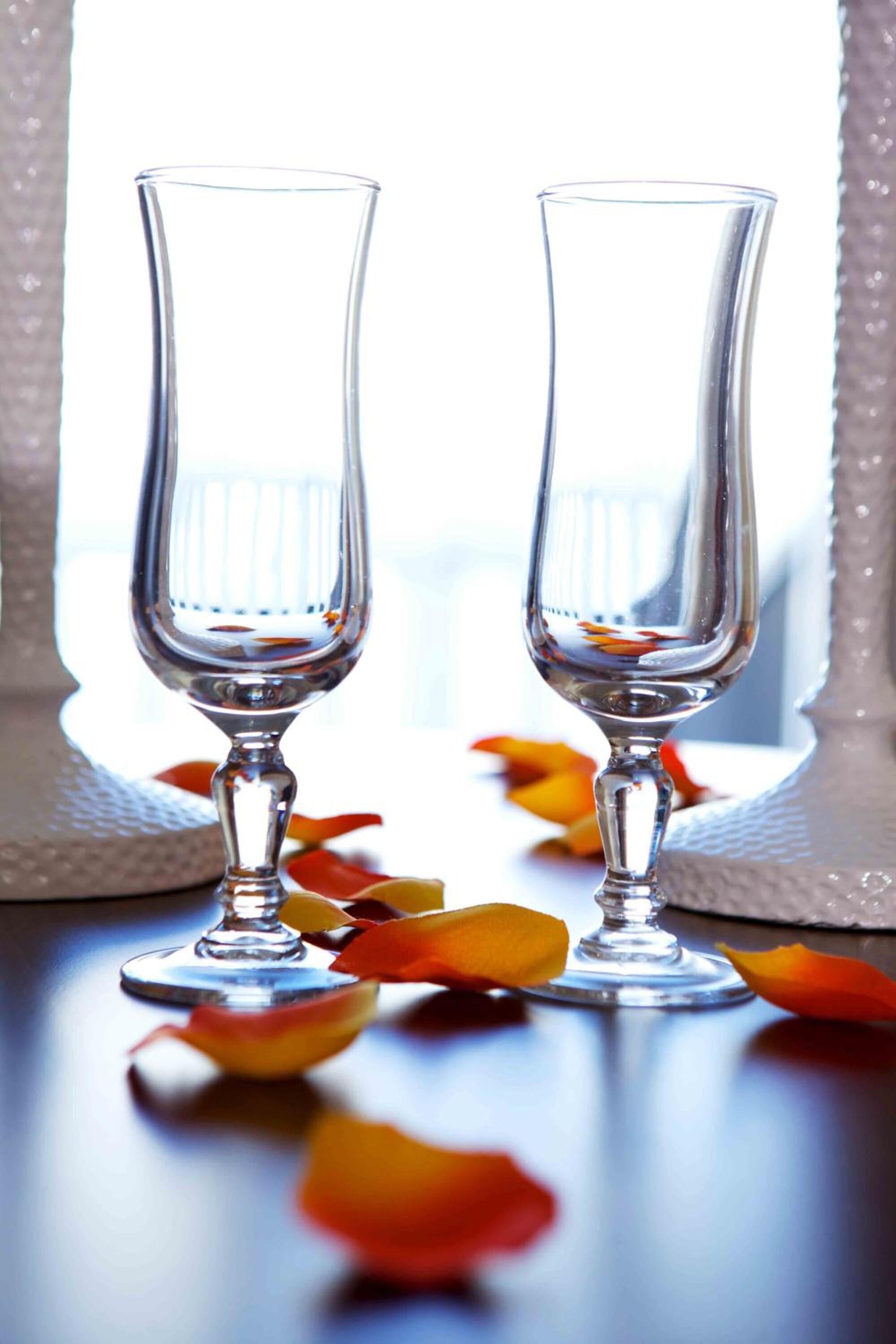 Two clear glass wine glasses on a reflective surface with scattered rose petals.