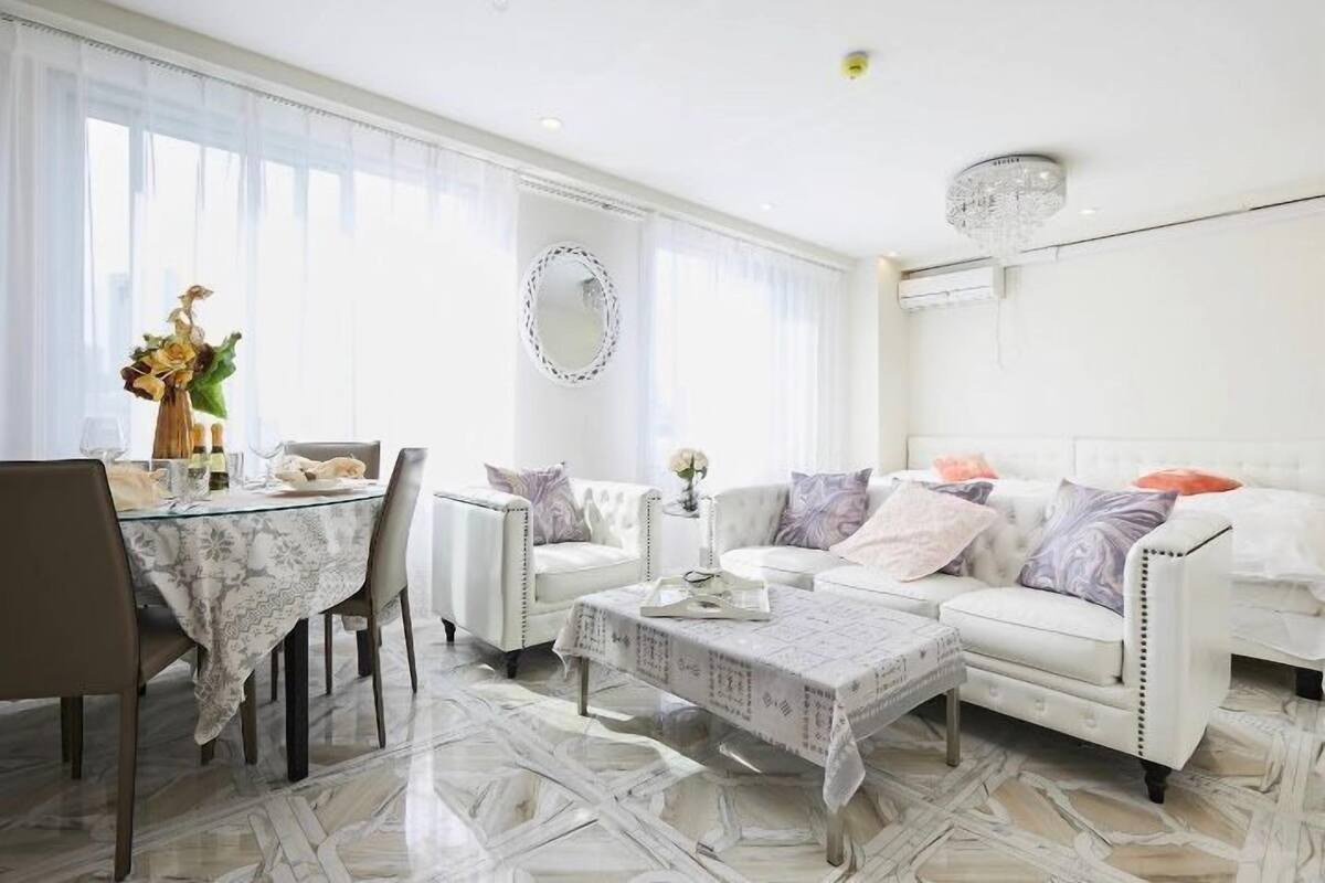 A modern living room with a white sofa, a glass coffee table, and a dining area with a table set for two.