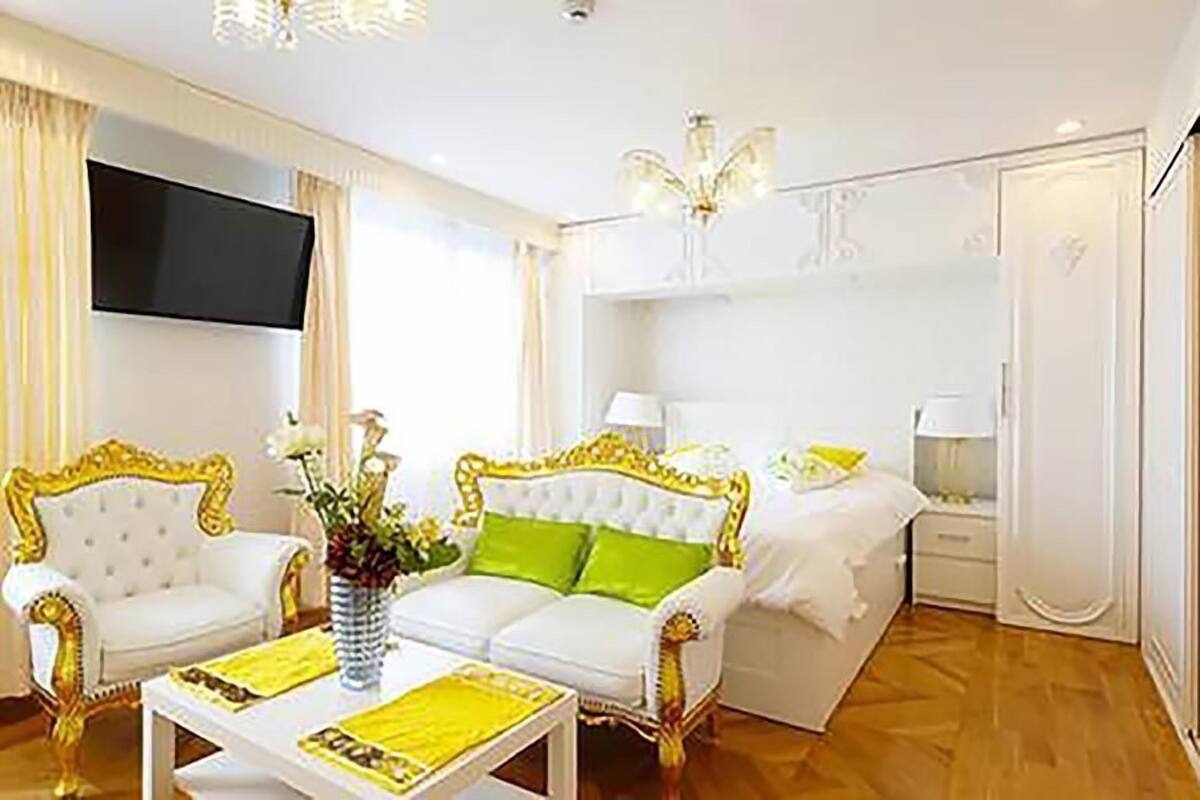 A hotel room with a white and gold sofa set, a bed with white bedding, a flat-screen TV mounted on the wall, and a chandelier.