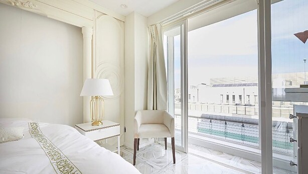 A hotel room with a large bed, a chair, a nightstand with a lamp, and a view of the city through large windows. - Reimi Mondo (Tokyo)