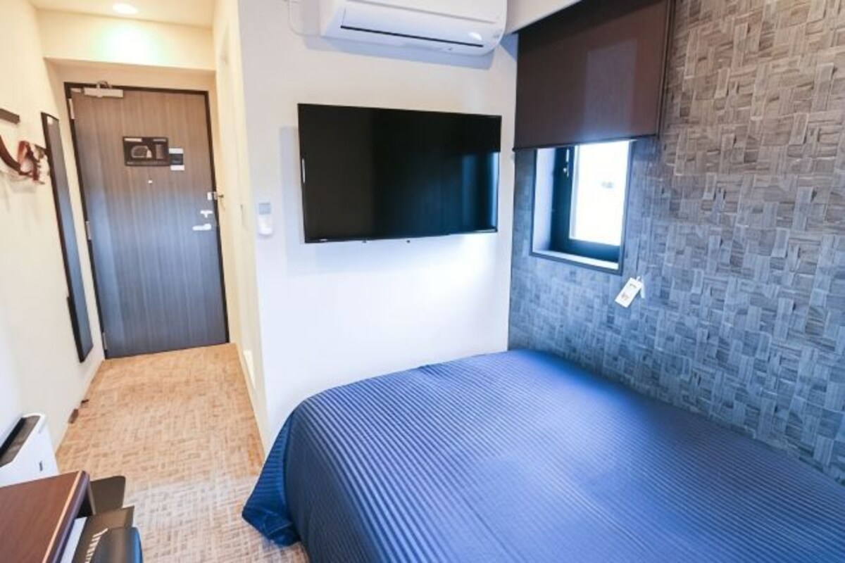 A bedroom with a bed, a TV mounted on the wall, a window, and an air conditioning unit.
