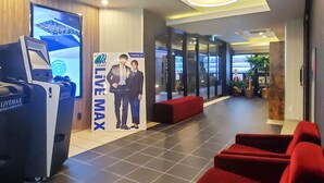 Zona de recepción de hotel con quiosco digital, cartel 'Live Max', asientos rojos y diseño de interiores moderno.