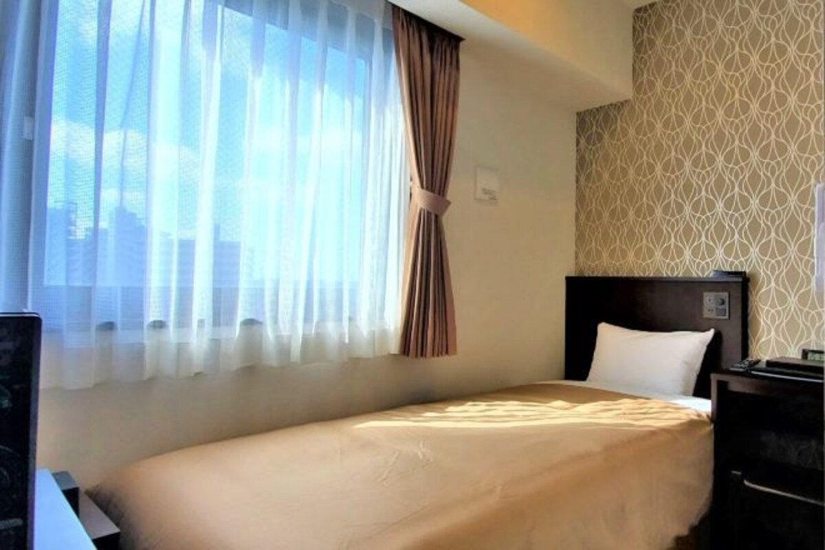 A hotel room with a bed, a headboard, a nightstand, and a window with sheer curtains.