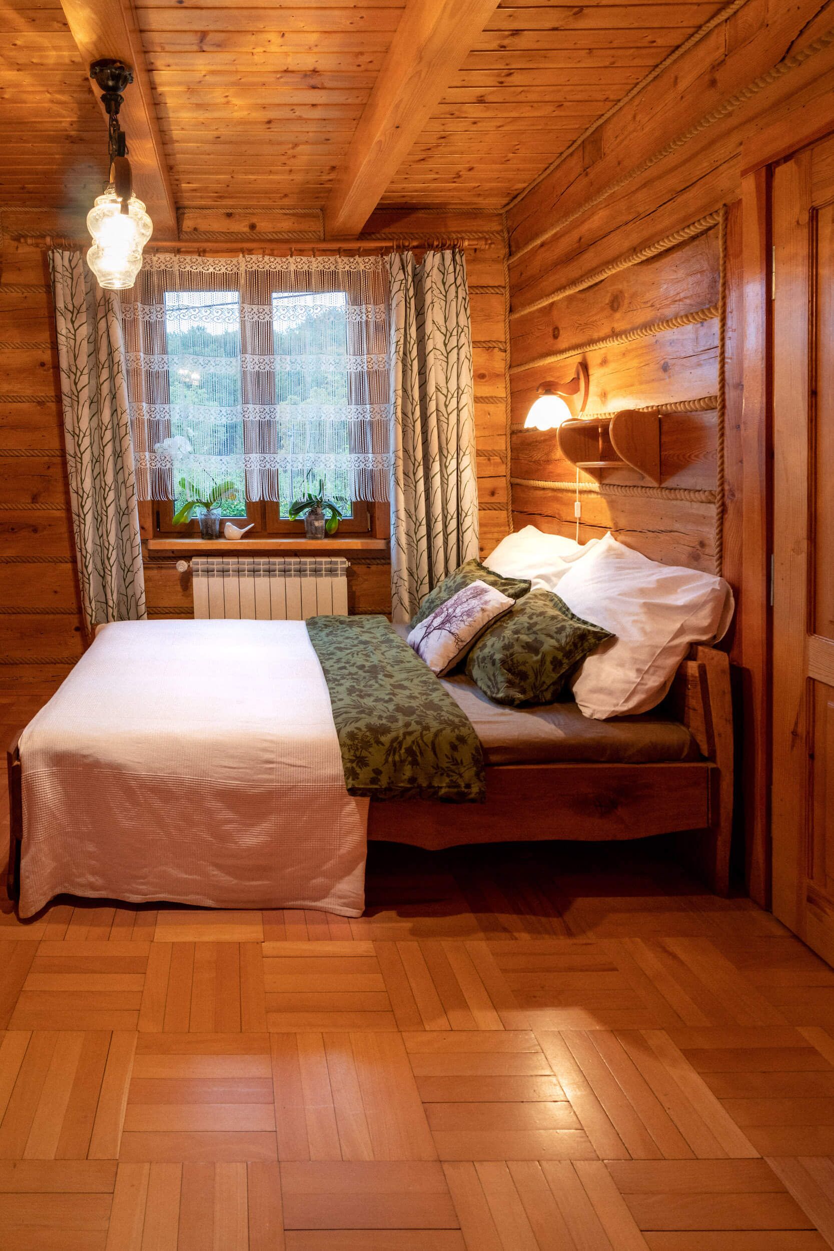 A wooden cabin bedroom with a bed, a window with curtains, a radiator, and a wall-mounted light fixture.