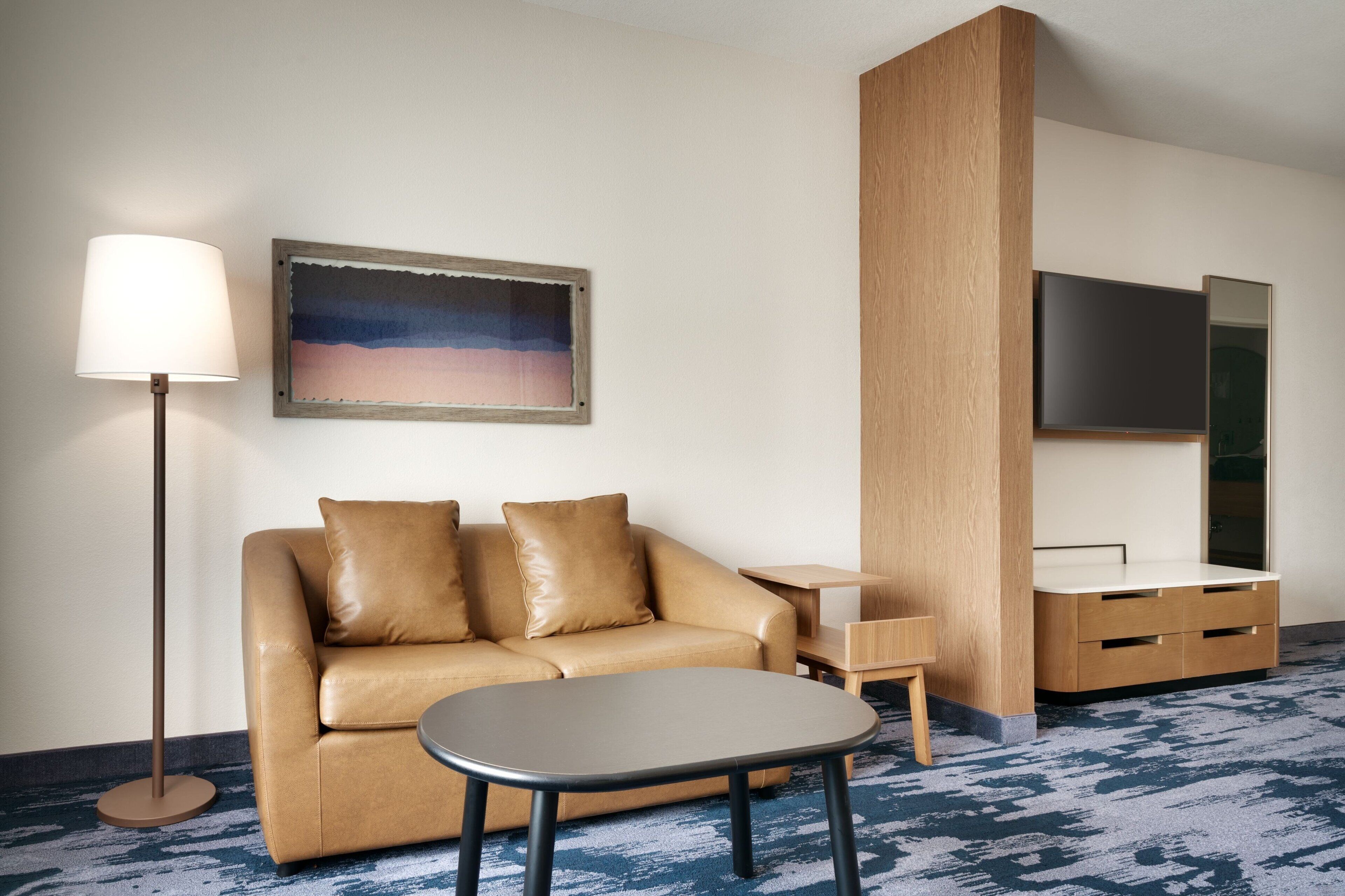 A hotel room with a sofa, a small table, a lamp, a painting, a TV, and a wooden cabinet.