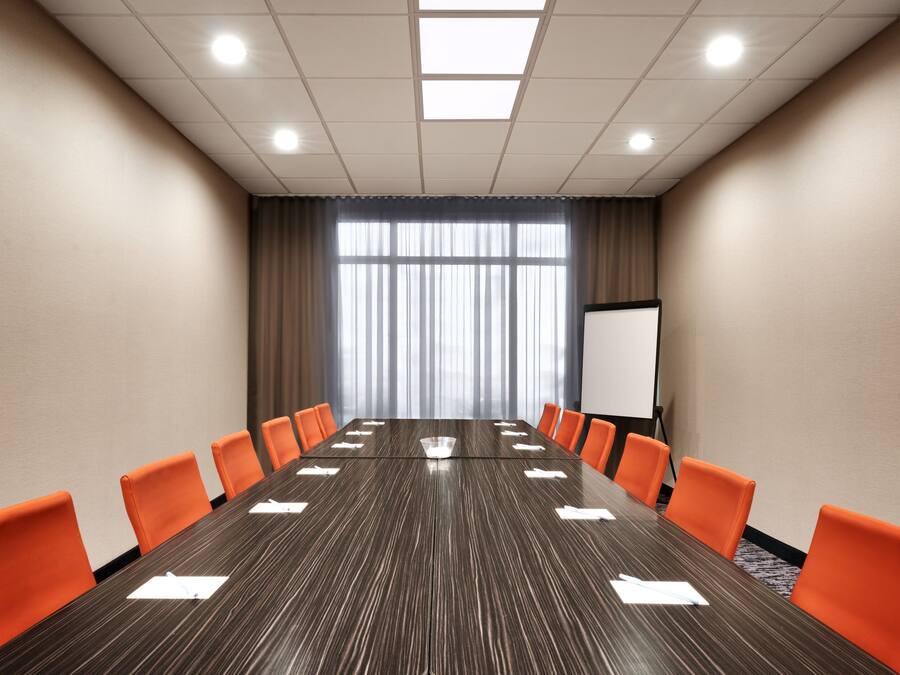 A conference room with a long table, orange chairs, a whiteboard, and a large window with curtains.