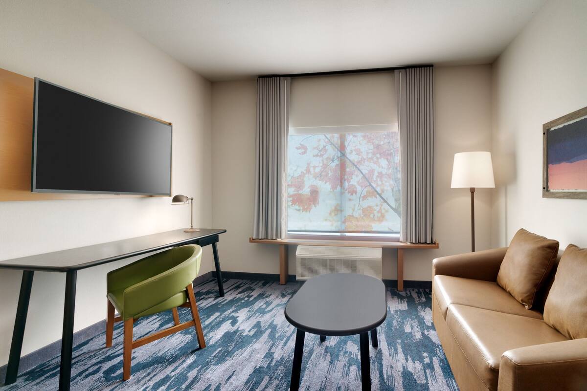 Habitación de hotel con televisor de pantalla plana, una silla verde, un sofá marrón, una mesita y una ventana con cortinas.