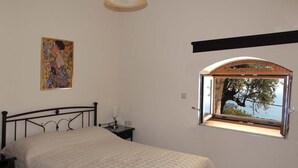 A bedroom with a bed, a nightstand, a lamp, a painting, and a window with a view of trees and the sea. - Olive Stonehouse Orestis (Kalamata)
