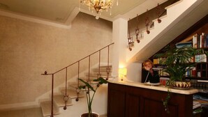 Zona de recepción con escalera, candelabro, planta en maceta y un escritorio con una persona detrás.
