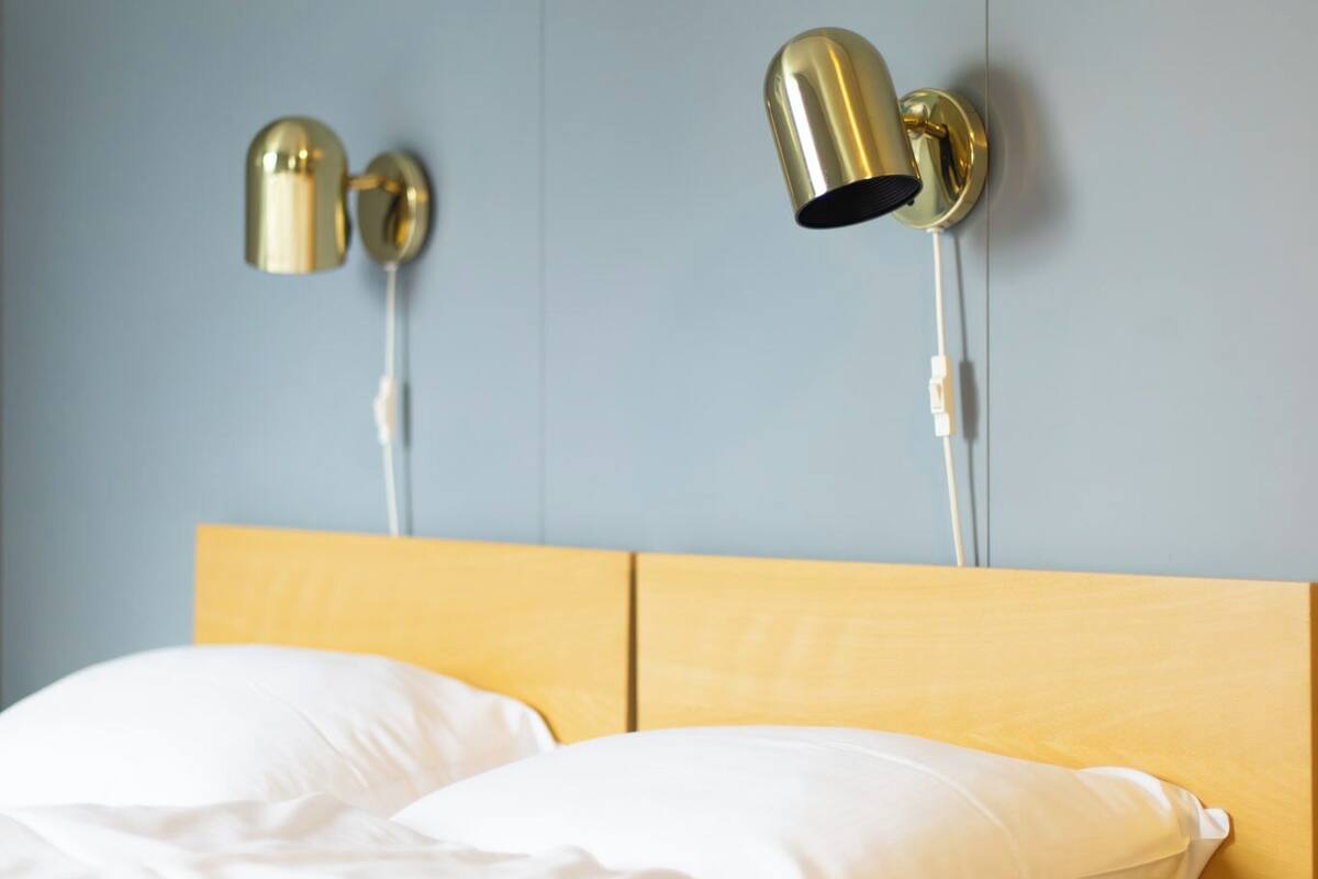 Una cama con ropa blanca y un cabecero de madera, dos lámparas fijadas a la pared y una pared lisa.