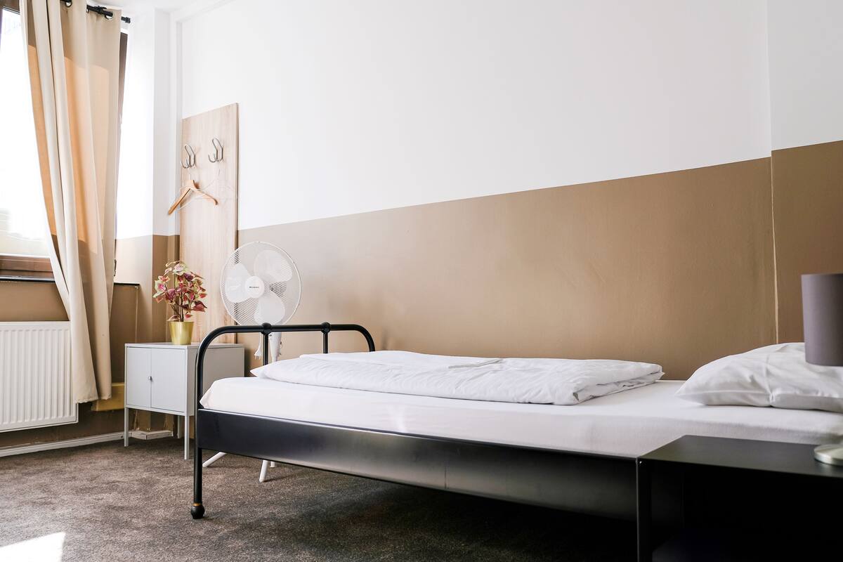 Ein ordentlich bezogenes Bett mit weißen Leinen, ein Nachttisch mit Ventilator, ein kleiner Schrank und ein Wandhaken für Kleidung.