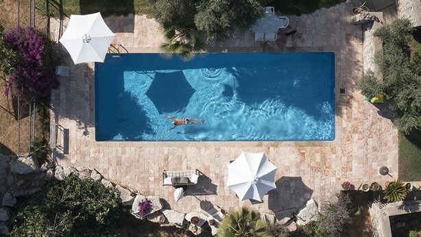 Une vue aérienne d’une piscine rectangulaire où une personne nage, entourée de meubles d’extérieur et de parasols, le tout aménagé dans un jardin paysager avec des arbres et des arbustes.